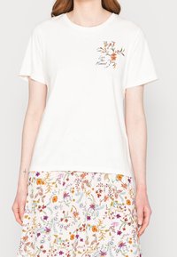 Camiseta de algodón blanca de mangas cortas, con un diseño floral y el texto "Vive el Momento" impreso en marrón oscuro.