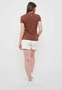 Maglietta a maniche corte marrone con scollatura rotonda e un piccolo logo sulla schiena, abbinata a pantaloni corti bianchi. Composizione in misto cotone, texture liscia.