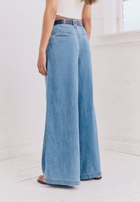 Leveälahkeiset vaaleansiniset farkut, joissa on tumma vyötärönauha, sileä pinta ja hillityt ompelutiedot, nähtynä takaa.