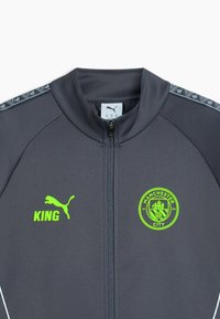 Tmavomodrošedá bunda na zips s neónovo zeleným logom Puma a textom "KING" na ľavej strane, erb Manchester City na pravej strane a vzorované pruhy na ramenách.