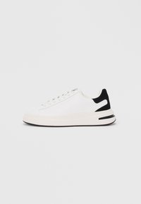 Baskets blanches avec un upper en cuir lisse, accent de talon en suède noir, lacets plats et semelle en caoutchouc texturée. Détail de logo notable sur le côté.