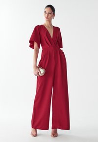 Rode jumpsuit met een diepe V-hals, brede mouwen en zijzakken. Gemaakt van gladde stof, heeft een getailleerde pasvorm en wijde benen.