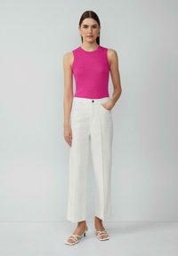 Haut sans manches rose ajusté associé à un pantalon blanc taille haute ; le mannequin se tient avec une main dans la poche ; design minimaliste et lignes épurées.