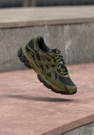 Deportiva deportiva de color verde oscuro y negro en el aire sobre una superficie de hormigón rosa con un muro de piedra gris de fondo.