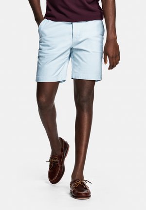 Man wearing light blue shorts and dark brown boat shoes, standing with hands in pockets against a plain background.