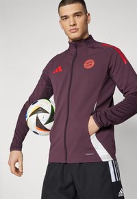 Bayern Miunchen zip-up striukė tamsiai burbulinėmis spalvomis su raudonais akcentais, baltais šonų skydeliais ir išsiuvinėtu logotipu, dėvima su futbolo kamuoliu.