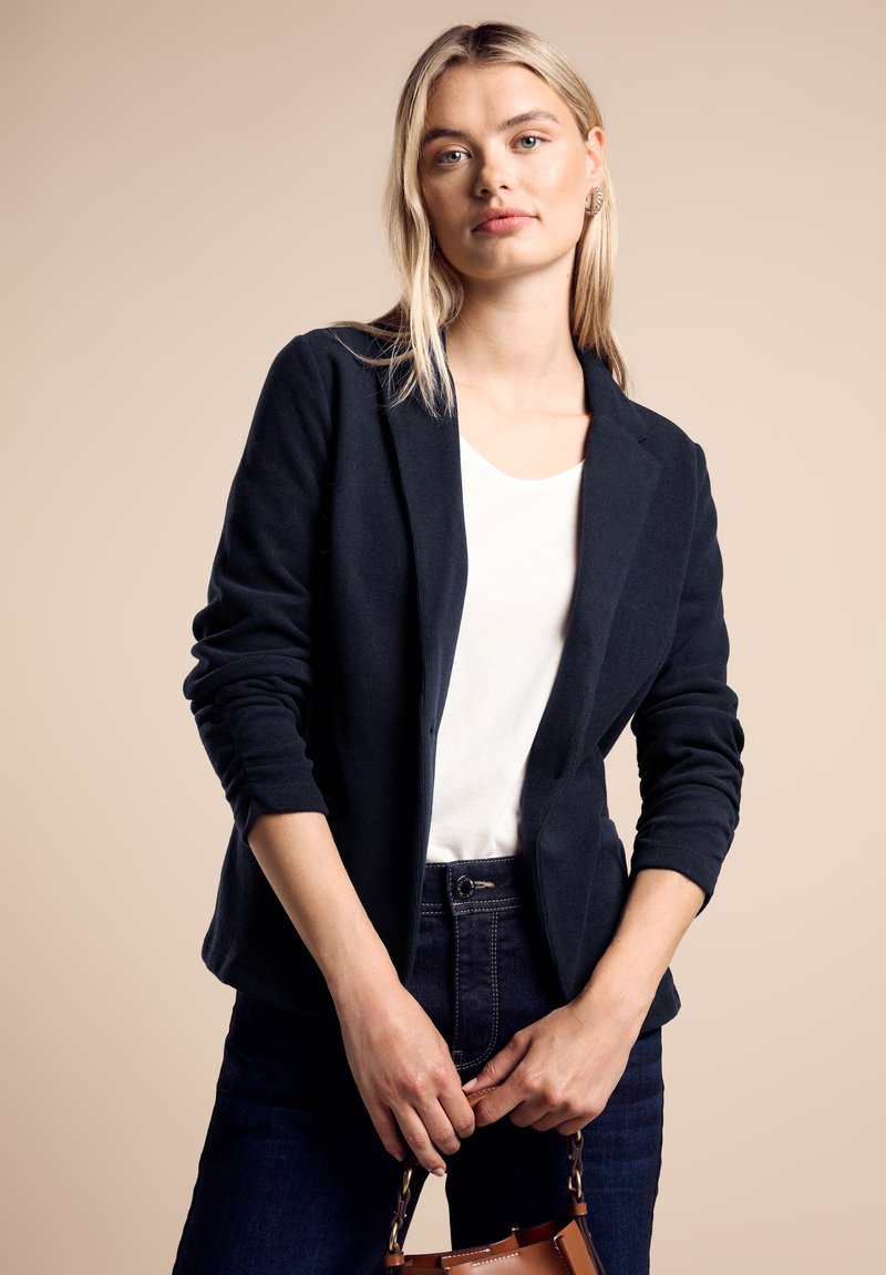 Blazer bleu marine en tissu doux, avec un col classique, une ouverture devant, et des manches longues, associé à un haut blanc et un jean sombre.