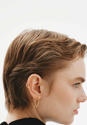 Pendientes de alambre dorado en formas abstractas. Uno es un corazón, el otro es un diseño curvado. Se ve el cabello corto y la piel clara de perfil.