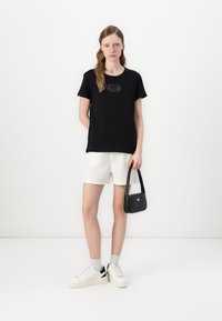Maglietta nera in cotone con un design grafico, abbinata a pantaloni corti bianchi in lino e scarpe da ginnastica bianche con un accent nero. Tiene in mano una piccola borsa nera.
