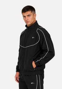 Jeune homme portant une veste de survêtement noire et un pantalon avec des passepoils blancs et un logo, debout avec une main dans la poche sur un fond uni.