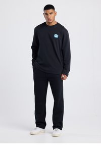 Black long-sleeve shirt with blue graphic detail on the chest, paired with black pants and white sneakers, set against a neutral background.