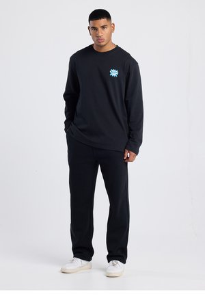 Schwarzes Langarmshirt mit blauem Grafikdetail auf der Brust, kombiniert mit schwarzer Hose und weißen Turnschuhen, vor einem neutralen Hintergrund.