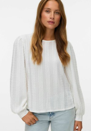 Vrouw met lang lichtbruin haar, gekleed in een witte textuur met lange mouwen blouse en lichtblauwe spijkerbroek tegen een effen achtergrond.