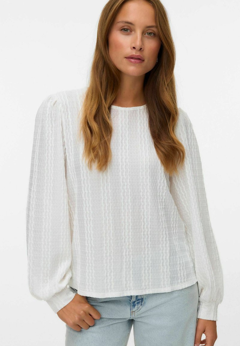 Femme avec de longs cheveux châtain clair, portant un chemisier blanc texturé à manches longues et un jean bleu clair, sur un fond uni.