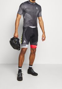 Cyklistické oblečení s černo-bílým pruhovaným krátkým rukávem a zipem, spárované s černými šortkami s vycpávkami a červenými a žlutými akcenty.