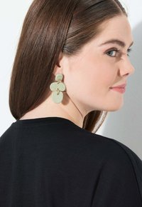Mujer con cabello castaño lacio que lleva unos grandes pendientes abstractos de color verde pálido y una blusa negra, mirando por encima de su hombro.