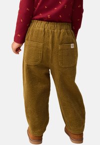 Pantalon en velours côtelé marron à jambe large, doté de deux poches arrière et d'une taille élastique, porté avec une chemise à manches longues à motifs rouges.