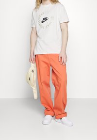 T-shirt Nike gris clair avec logo noir, associé à un pantalon orange ample et des baskets blanches, tenant un sac jaune.