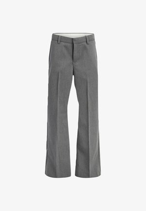 Gutt som står iført en olivengrønn skjorte med knapper, grå bukser og beige sko mot en ensfarget lys bakgrunn.