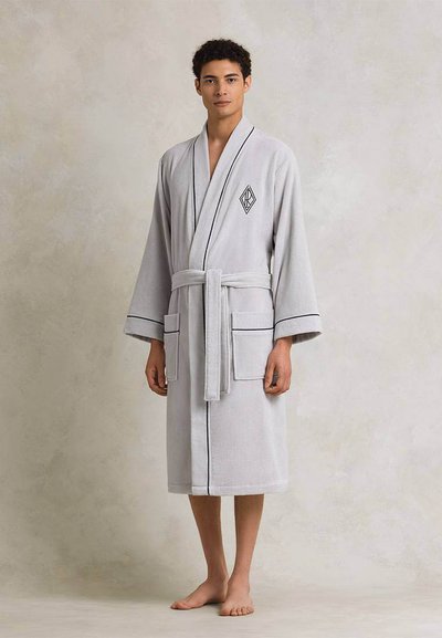 Νεαρός άνδρας που στέκεται ξυπόλητος φορώντας ένα ανοιχτό γκρι μπουρνούζι με μαύρη λεπτομέρεια και ζώνη, μπροστά σε ουδέτερο φόντο.