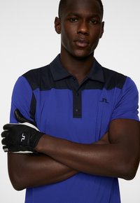 Blaues Poloshirt mit dunklem Akzent, drei Knöpfen und Logo. Schwarze Golfschaufel mit weißen Details an der linken Hand, Arme gekreuzt. Glatte Textur.