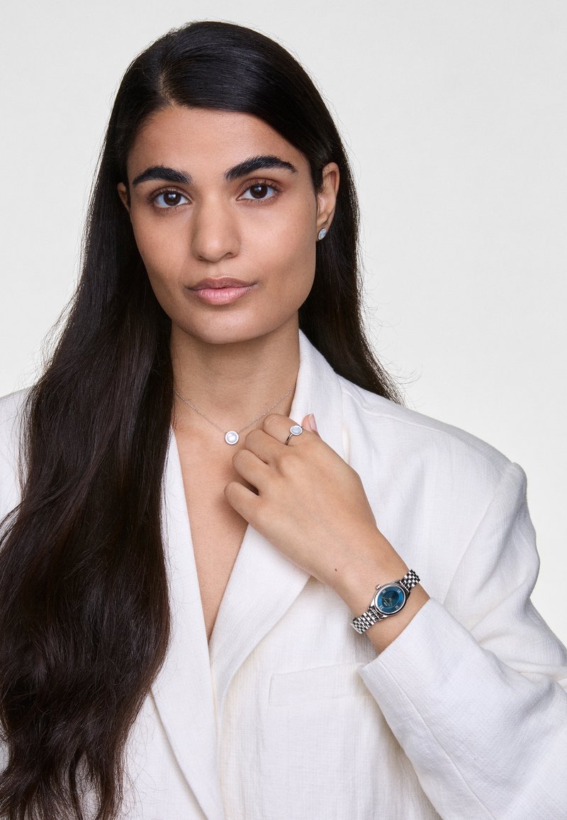 Silver watch with a blue face on a mesh bracelet. Woman wearing a necklace with a circular pendant and a matching ring. Light-colored blazer.