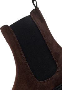 Botín de ante marrón con un panel lateral elástico negro. Presenta una costura a lo largo del borde y una lengüeta para facilitar su colocación.