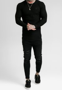 Schwarzes, geripptes Langarmshirt mit Logo, kombiniert mit schwarzen, zerrissenen Skinny Jeans und schwarzen Sneakers mit goldenen Akzenten.