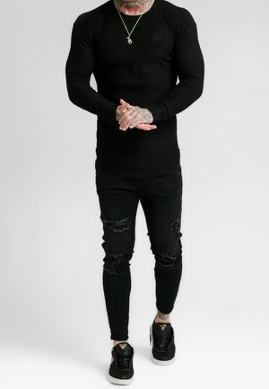 Black ribbed long-sleeve shirt with logo, paired with black ripped skinny jeans and black sneakers featuring gold accents.