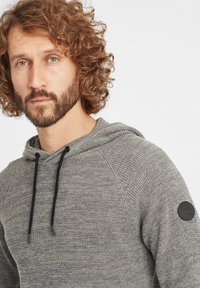 Szara bluza z kapturem, wykonana z teksturowanej tkaniny, z kapturem na sznurku, posiadająca czarną naszywkę z logo na lewym ramieniu oraz prążkowe detale.