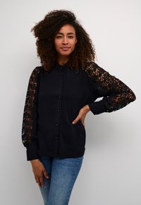 Black button-up shirt with intricate black lace sleeves, featuring a collar and a relaxed fit, paired with blue jeans.