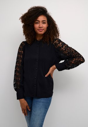 Black button-up shirt with intricate black lace sleeves, featuring a collar and a relaxed fit, paired with blue jeans.