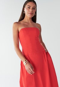 Punane rihmadeta kleit, millel on vormiv ülakeha ja voolav seelik. Valmistatud kergest kangast, millel on sile tekstuur ja peened õmblused.