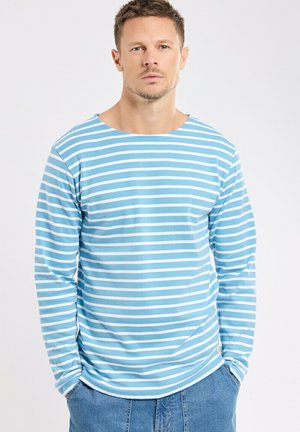 Homme aux cheveux courts portant une chemise à manches longues à rayures bleu clair et blanc et un jean bleu, debout les mains dans les poches.