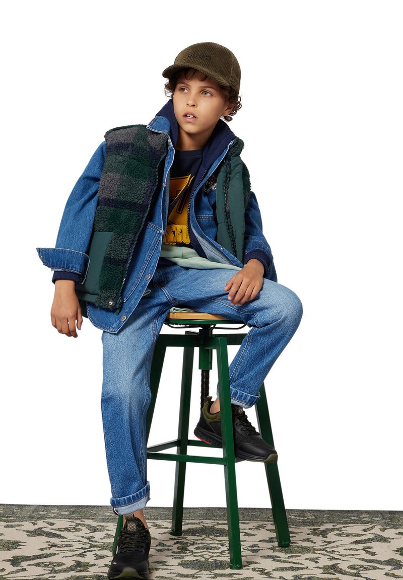 Veste en jean superposée sur un gilet à carreaux vert et noir, assortie à un jean bleu et des chaussures noires. Une casquette cordonnée verte complète la tenue.