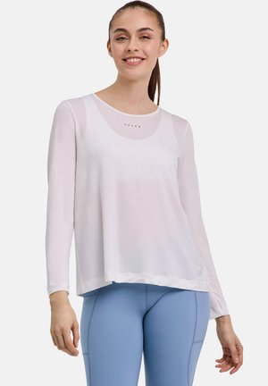 Donna sorridente con capelli castani raccolti in una coda di cavallo indossa una maglia bianca a maniche lunghe trasparente e leggings azzurri chiari su uno sfondo uniforme.