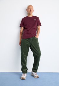 Maroon cotton t-shirt with a small white logo, paired with green corduroy pants, and white sneakers featuring gray and brown accents.