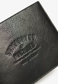 Cartera de cuero negro con superficie texturada, logotipo en relieve "SUPERDRY CO. MACHINED" y costuras cuidadas en los bordes.