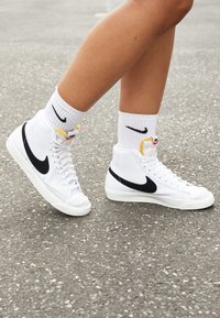 Baskets montantes blanches en matériau synthétique, avec un swoosh noir de Nike, un accent jaune et une semelle en caoutchouc blanc texturé.