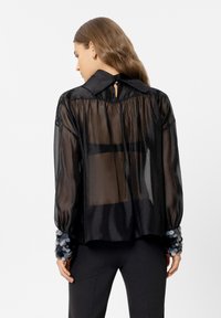 Schwarze, transparente Bluse mit einem knöpfbaren Kragen, puffartigen langen Ärmeln und mit Pailletten verzierten Bündchen in Grautönen. Detail mit Schlitz auf der Rückseite.
