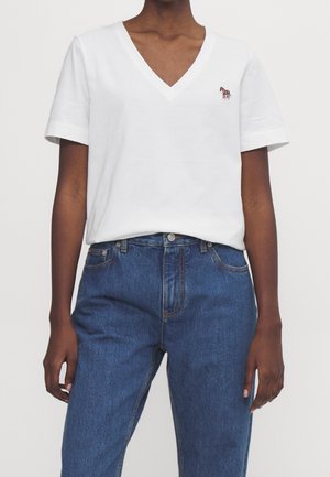 Personne portant un t-shirt blanc à col en V avec un petit logo zèbre sur la poitrine et un jean bleu taille haute.