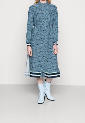 Niebieska sukienka w wzory z długimi rękawami, zapinana na guziki z przodu, z taliowaniem i kontrastującymi mankietami, zestawiona z jasnoniebieskimi botkami na obcasie.