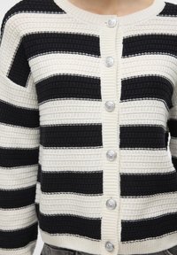 Stickad cardigan med svarta och gräddvita horisontella ränder, med en texturerad kostym och fyra silverknappar framtill.