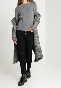 Grå sweater med en tekstureret overflade, sorte jeans med tre knapdetaljer, og en lang ternet frakke i jordtoner, sat sammen med sorte støvler.