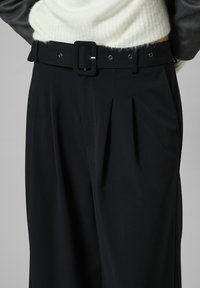 Fekete, magas derekú nadrág pliszékkal, széles övvel, négyzet alakú csattal és fém szemekkel. Puha szövet szerkezet, szabott kialakítás.