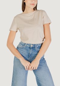 T-shirt in cotone beige con maniche corte, caratterizzato da un logo "GUESS" in rilievo sul petto, abbinato a jeans di denim azzurro chiaro.