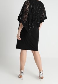 Frau, die einen schwarzen Spitzen-Bolero mit Pailletten, einen schwarzen knielangen Rock und silberne High-Heels trägt, steht vor einem schlichten Hintergrund.