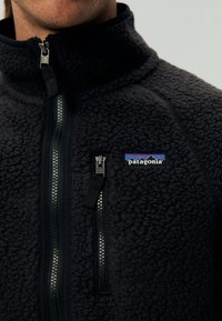 Musta fleecekangas takki korkean kauluksen kanssa, jossa on vetoketjusuljenta ja rintatasku. Patagonia-logo vasemmalla puolella. Rakenteellinen kangas.