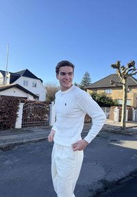 Tânăr îmbrăcat în pulover alb și pantaloni, stând pe o stradă rezidențială cu case, un copac fără frunze și cer albastru clar în fundal.