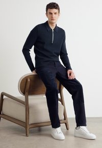 Marineblauwe longsleeve gebreide polo met een ritskraag, gecombineerd met donkere broeken en witte sneakers, zittend op een lichthouten stoel.